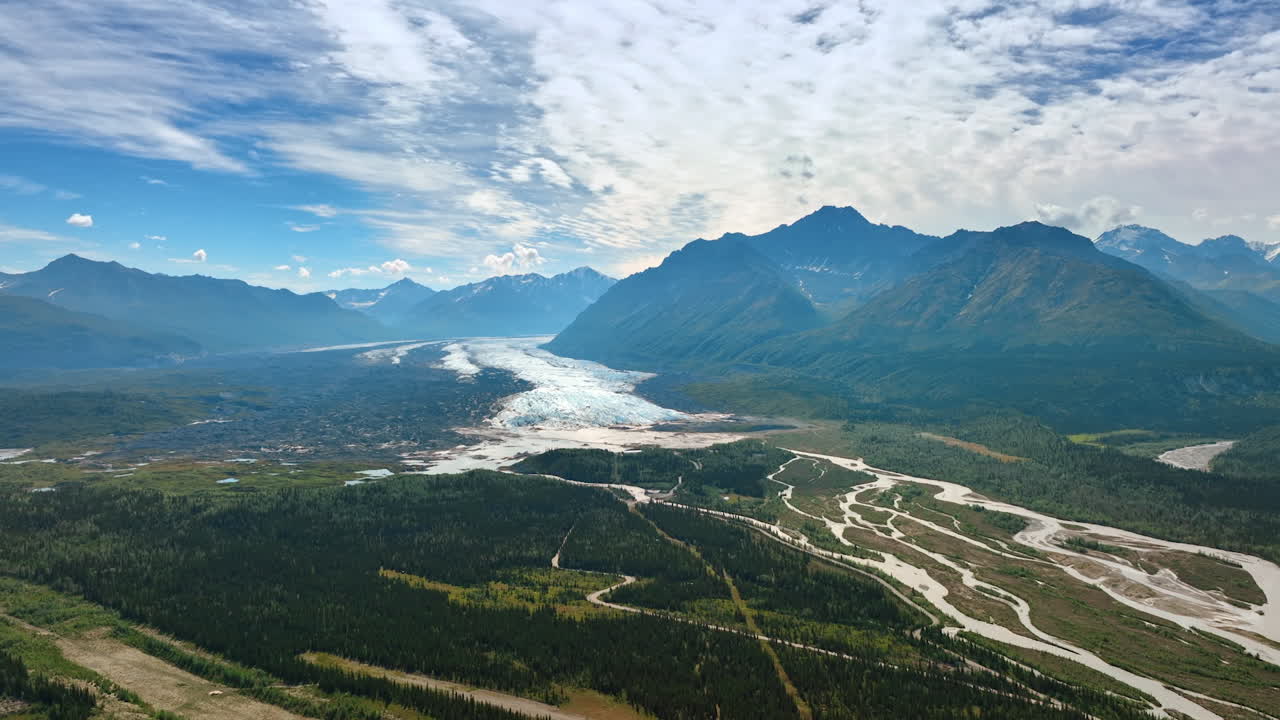 Striking panorama of mountains dazzled by the bright sun. White clouds cover the sky. Branching river flows in the valley. Aerial view. Alaska, USA