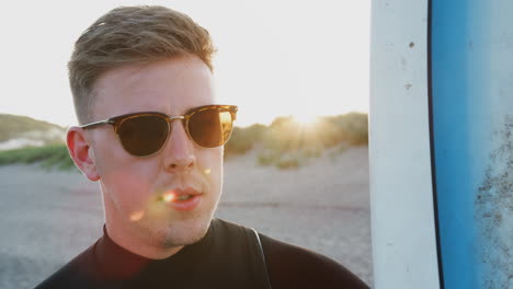 young man in sunglasses wearing wetsuit enjoying surfing vacation on beach as sun sets