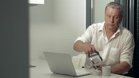 hombre adulto serio trabajando en la computadora portátil mientras bebe café en la cocina