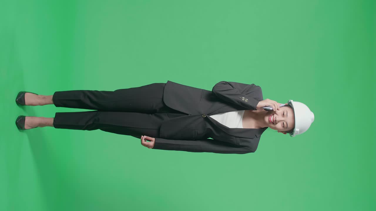 Full Body Of Smiling Asian Female Engineer With Safety Helmet Talking On Smartphone In The Green Screen Studio