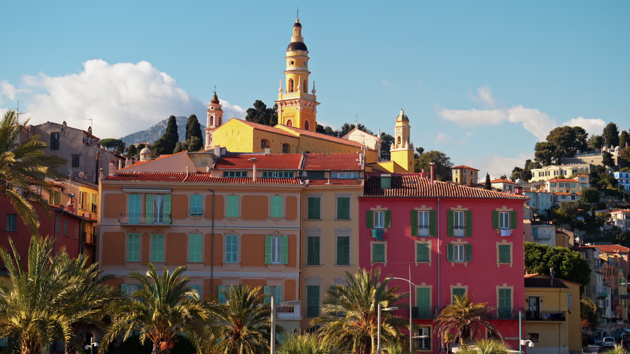 Distant view of the St Michel Basilica surrounded by colourful buildings and palm trees, Menton, France