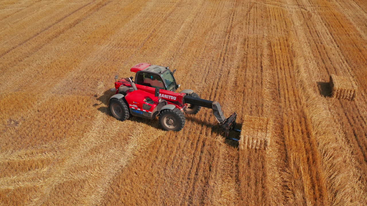 Little agile powerful loader machine picks up hay bale and puts it on another one. Agricultural machinery working in the field after harvesting wheat.