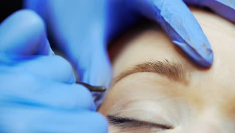 Tattoo specialist in gloves is drawing eyebrows with a thin needle for a woman in a beauty salon. Close-up