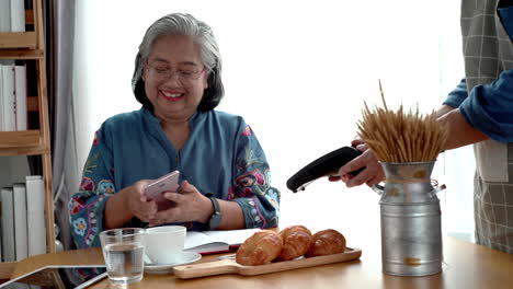 Active-senior-Asian-woman-working-in-a-coffeeshop-using-a-mobile-device,-smartwatch-for-NFC,-QR-code-scanning,-contactless-payment-to-pay-for-coffee-and-snack