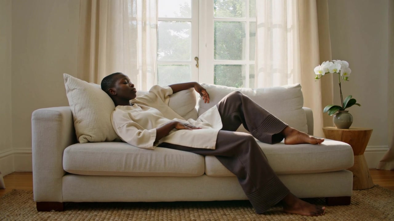 Reclining man shifting slightly for comfort on light sofa cushion in living room, wearing cream top