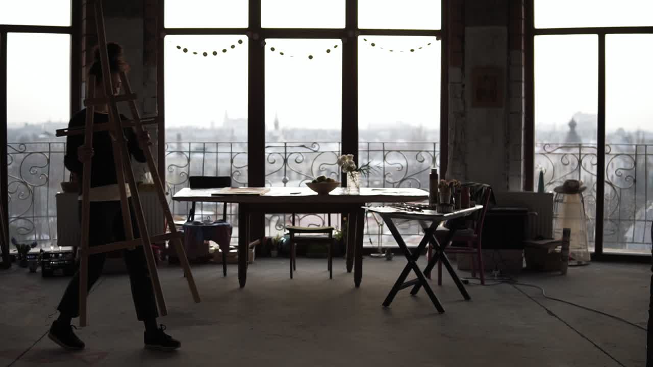 Female artist, shaded by the day light on the bakground coming from panoramic windows, brings in the easel, puts canvas on it and starts drawing. Slow motion footage.