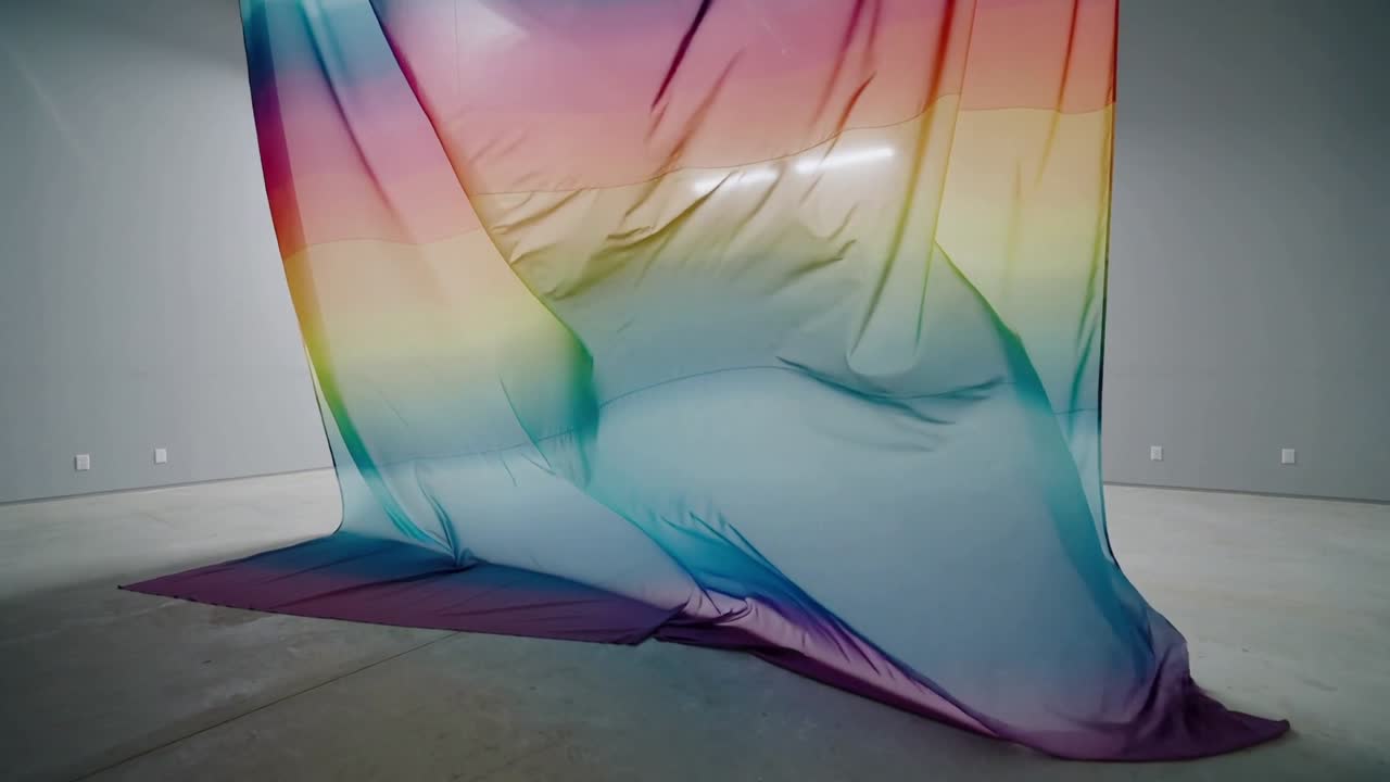 Tessuto arcobaleno che ondeggia in galleria mentre l'aria muove le pieghe sotto una luce dall'alto mostrando una nuova forma