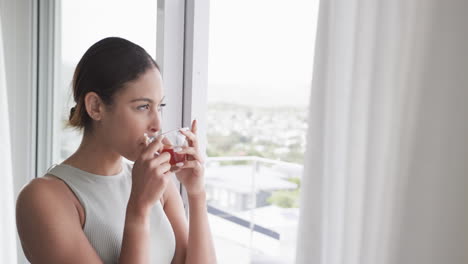 Mujer-Birracial-Pensativa-Mirando-Por-La-Ventana-Bebiendo-Té-En-Casa,-Copiando-Espacio,-Cámara-Lenta