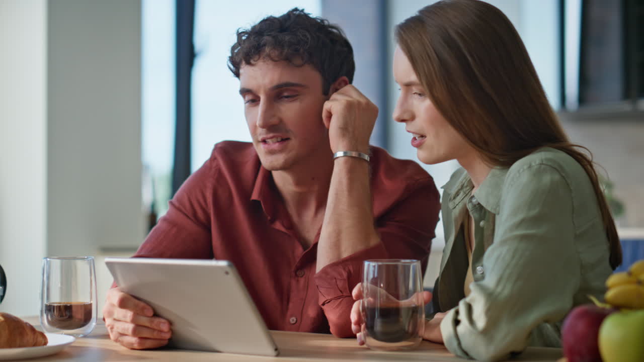 Smiling spouses holding tablet having breakfast at home closeup. Man watching