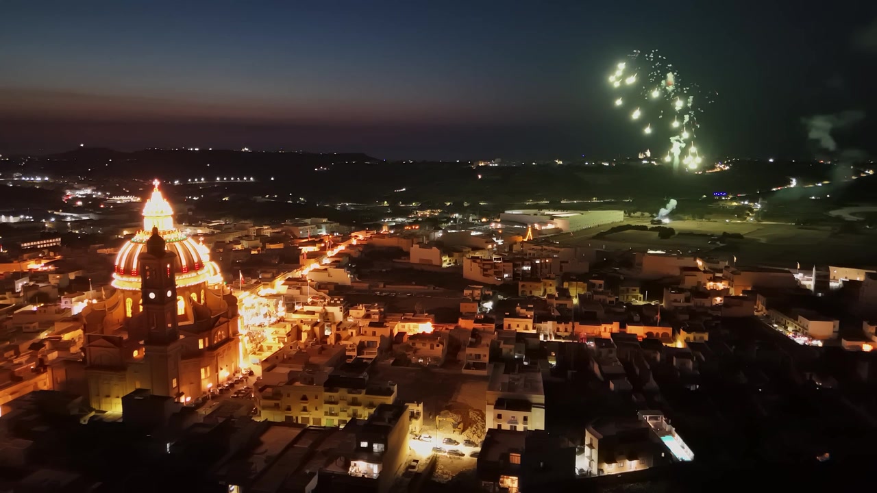 Festa del villaggio di Xewkija, Gozo, con fuochi d'artificio intorno alla Chiesa di San Giovanni Battista e strade illuminate al tramonto, con la cupola della basilica e il paesaggio urbano circostante visti da un drone