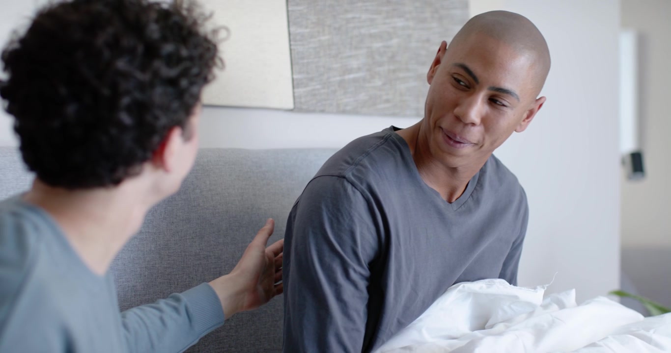 Mid 20s men sitting on bed, left man holding remote initiating right man waking, stretching