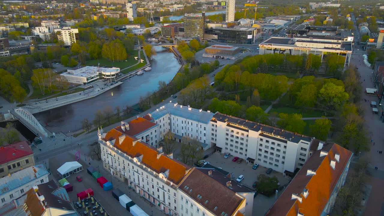 Beautiful aerial drone footage view of Tartu town hall square with cobblestone roads and historic heritage white medieval building during a sunny evening day while a large Emajõgi river flows in city