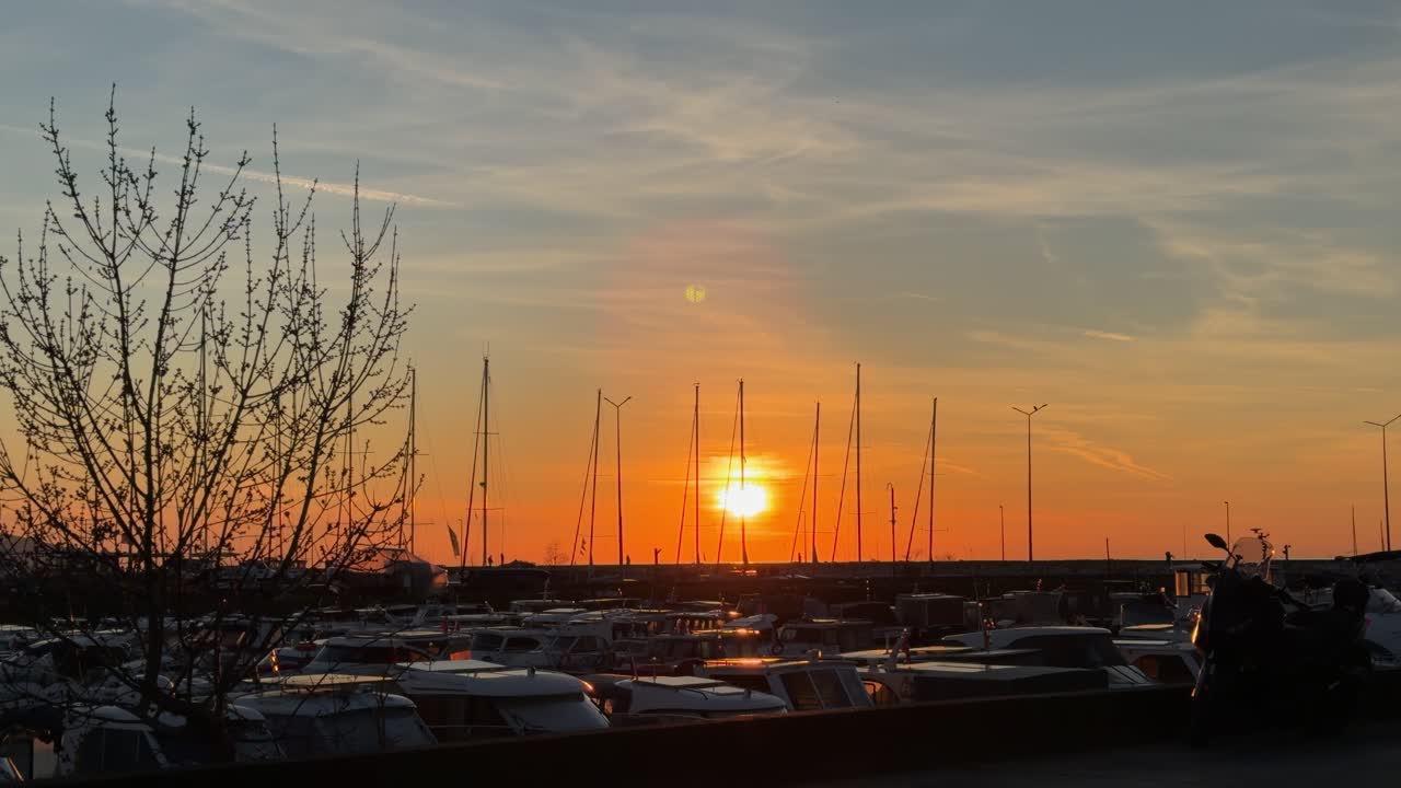Vakker gyllen solnedgangsutsikt i en fredelig havn med silhuetter av seilbåtmaster og parkerte båter. Folk går forbi og nyter den romantiske kveldsatmosfæren ved marinaen