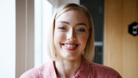 Young-white-woman-standing-by-the-window-in-the-office-laughing-to-camera,-head-and-shoulders,-close-up