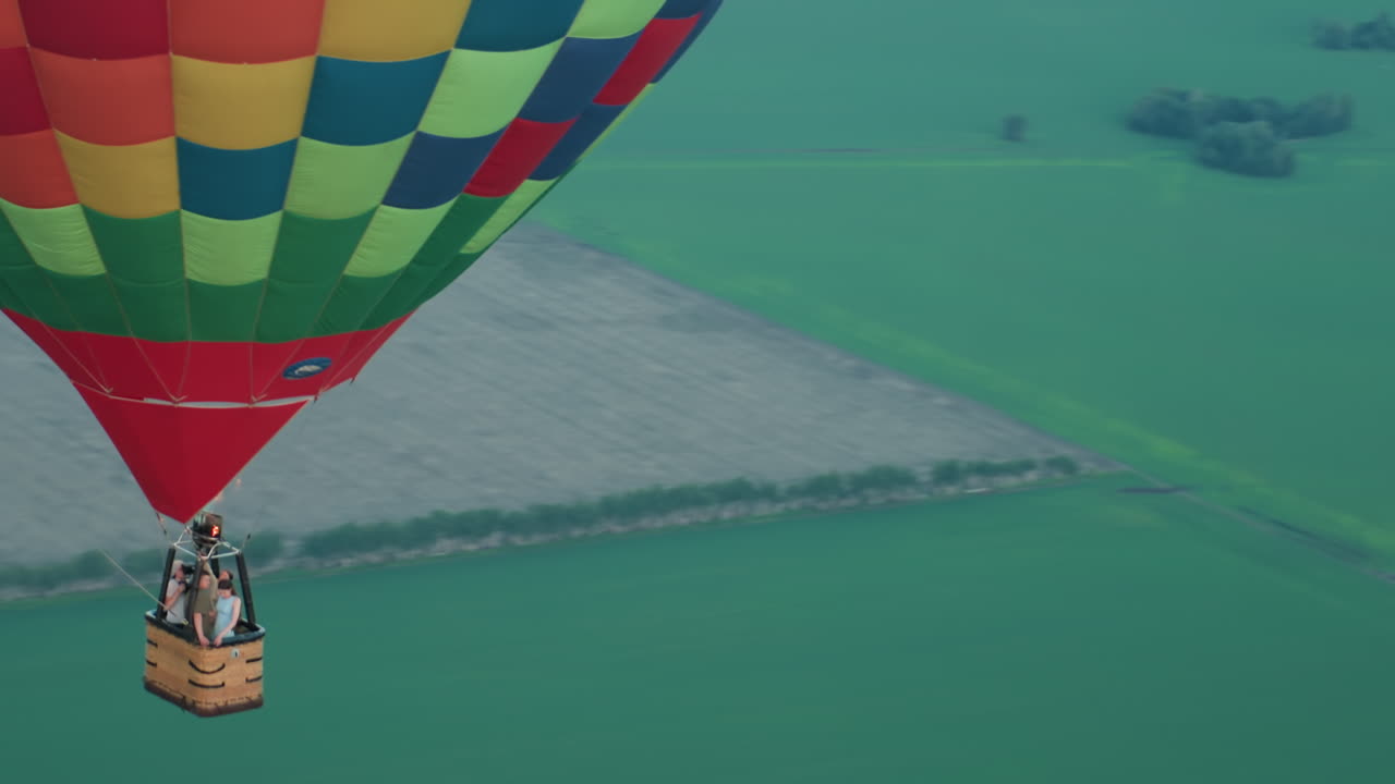low close up of colourful green red hot air balloon carrying small group of passengers in wicker basket gliding quietly over striped patchwork farmland at golden evening light