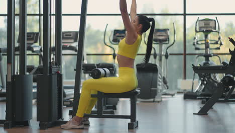 Una-Mujer-Hispana-Sentada-En-Un-Simulador-En-El-Gimnasio-Tira-De-Una-Cuerda-De-Metal-Con-El-Peso-Bombea-Los-Músculos-De-La-Espalda.-Mujer-Morena-Tira-Del-Simulador.-Realización-De-Ejercicios-Para-El-Simulador-De-Músculos-De-La-Espalda.