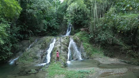 Pan-right-aerial-view-of-a-young-woman-standing-at-the-base-of-a-cascading-waterfall
