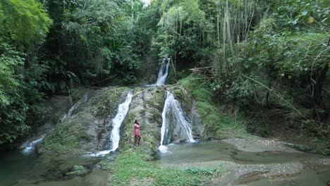 Schwenken-Sie-Nach-Rechts-Luftaufnahme-Einer-Jungen-Frau,-Die-Am-Fuß-Eines-Kaskadierenden-Wasserfalls-Steht