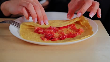 close-up, hands rolling homemade crepe with sweet strawberry jam on white plate