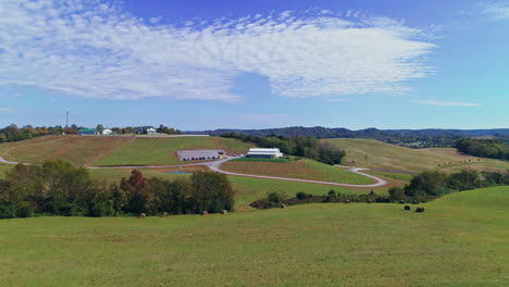 Aerial-passing-over-hay-bales-in-hilltop-to-reveal-barn-in-rural-farmland,-4K