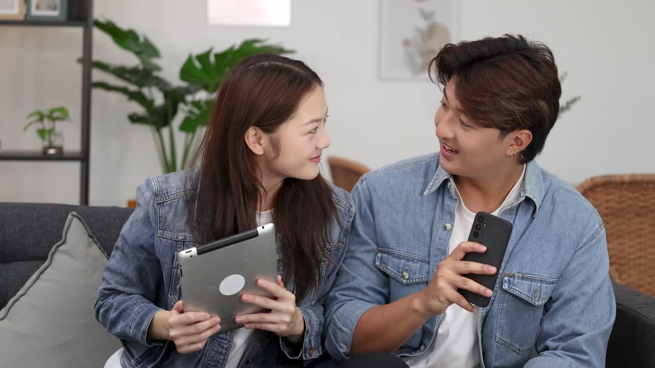 若いアジア人のカップルがソファーに座って、タブレットとスマートフォンを使いながら話しています。