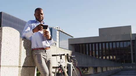 Afroamerikanischer-Mann-In-Der-Stadt-Mit-Fahrrad,-Der-In-Der-Sonne-Steht,-Kaffee-Trinkt-Und-Sein-Smartphone-Benutzt