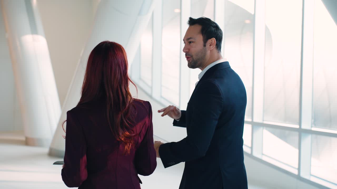 Plano de seguimiento trasero de un empresario y una empresaria caminando y conversando mientras revisan gráficos de informes coloridos en el vestíbulo de una oficina corporativa moderna y luminosa.