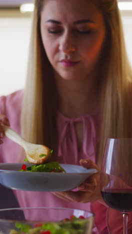 attractive long haired woman in elegant dress with wineglass of red wine takes delicious salad from glass bowl at romantic dinner at home slow motion