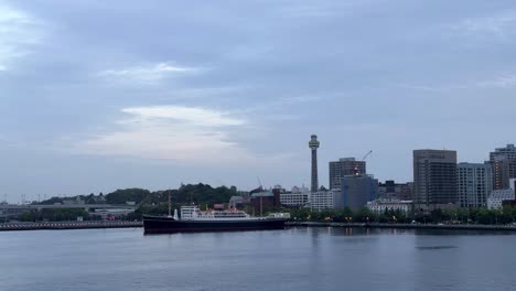 Dusk-settles-over-city-skyline-with-iconic-tower-and-moored-ship,-gentle-water-in-the-foreground