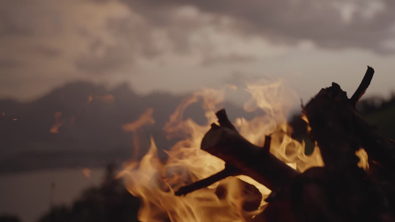 A campfire burns on a grassy hill with a mountain range and lake in the background under a dramatic evening sky. Concept of adventure, nature, solitude, and outdoor lifestyle