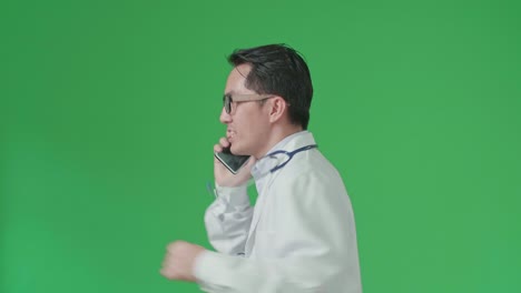 the side view of asian doctor with stethoscope wearing glasses and white coat talking on mobile phone while running on green screen chroma key.