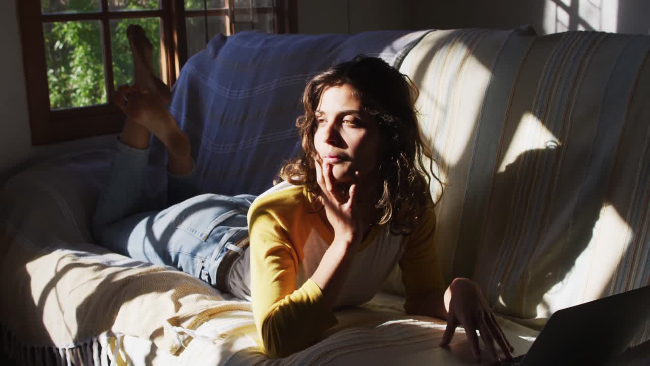 mujer de raza mixta pensativa acostada en el sofá usando una computadora portátil en la sala de estar soleada