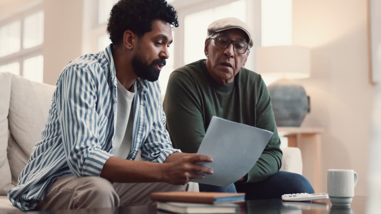 un hombre mayor y un hombre más joven se sientan en un sofá y miran un documento juntos.