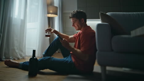 Relaxed man drinking cognac in night home interior. Laughing guy check sms alone