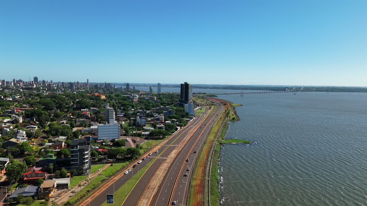 Højhastigheds-lufttur langs Costanera-motorvejen i Posadas, Misiones, med flere biler, der kører mellem byen og Paraná-floden under en lys, solrig morgen med blå himmel