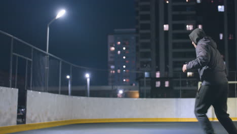 vista lateral de um homem de capuz chutando uma bola de futebol contra a parede sob iluminação brilhante do estádio à noite, paisagem urbana com edifícios iluminados e poste de gol no fundo