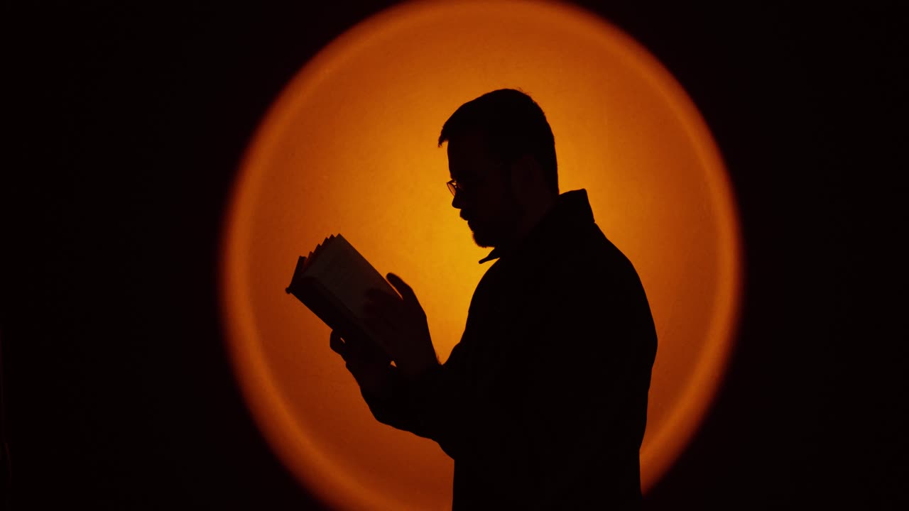 Silhouette cinematografica di profilo laterale di un uomo che legge un libro con una retroilluminazione circolare arancione incandescente in uno studio buio. Ideale per concetti come ricordi, riflessione, apprendimento, narrazione, mistero, a