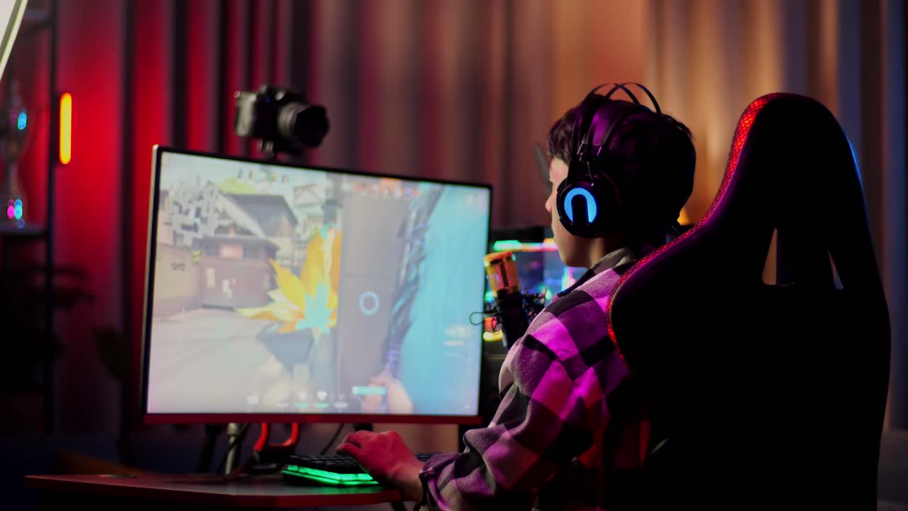 vista trasera de un chico asiático jugando a un juego en red en una computadora personal. videojuego de transmisión en vivo, escritorio iluminado por luz de tira led rgb