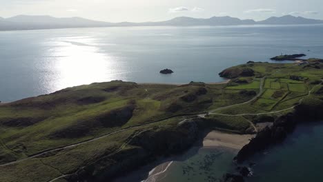 Vista-Aérea-Retroceda-A-Través-De-La-Isla-Ynys-Llanddwyn-Y-La-Brumosa-Cordillera-De-Snowdonia-Con-El-Brillante-Mar-Irlandés-Al-Amanecer.