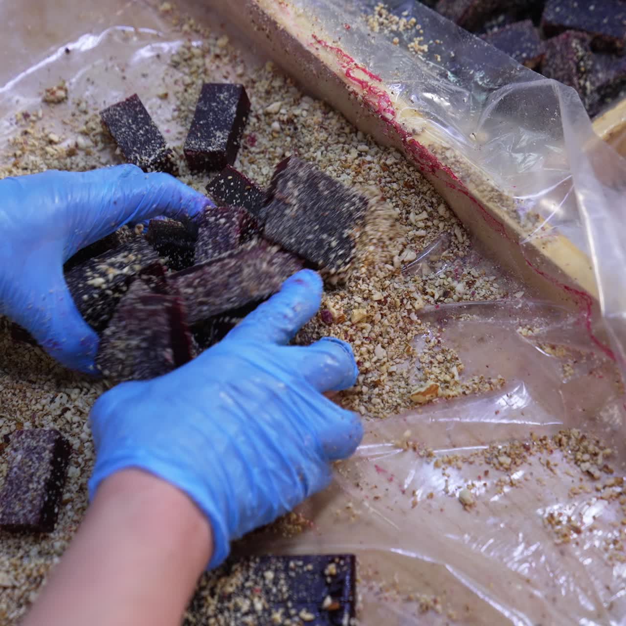 Dark marmalade pieces being coated with crushed nuts. Worker's hands mix up sweet desserts with ground walnuts. Close up