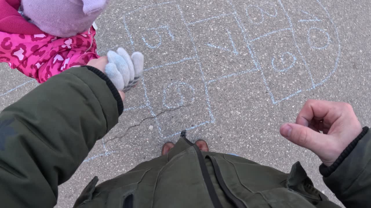 A heartwarming view from a parent’s perspective as they hold their child’s hand, watching them jump while playing hopscotch.
