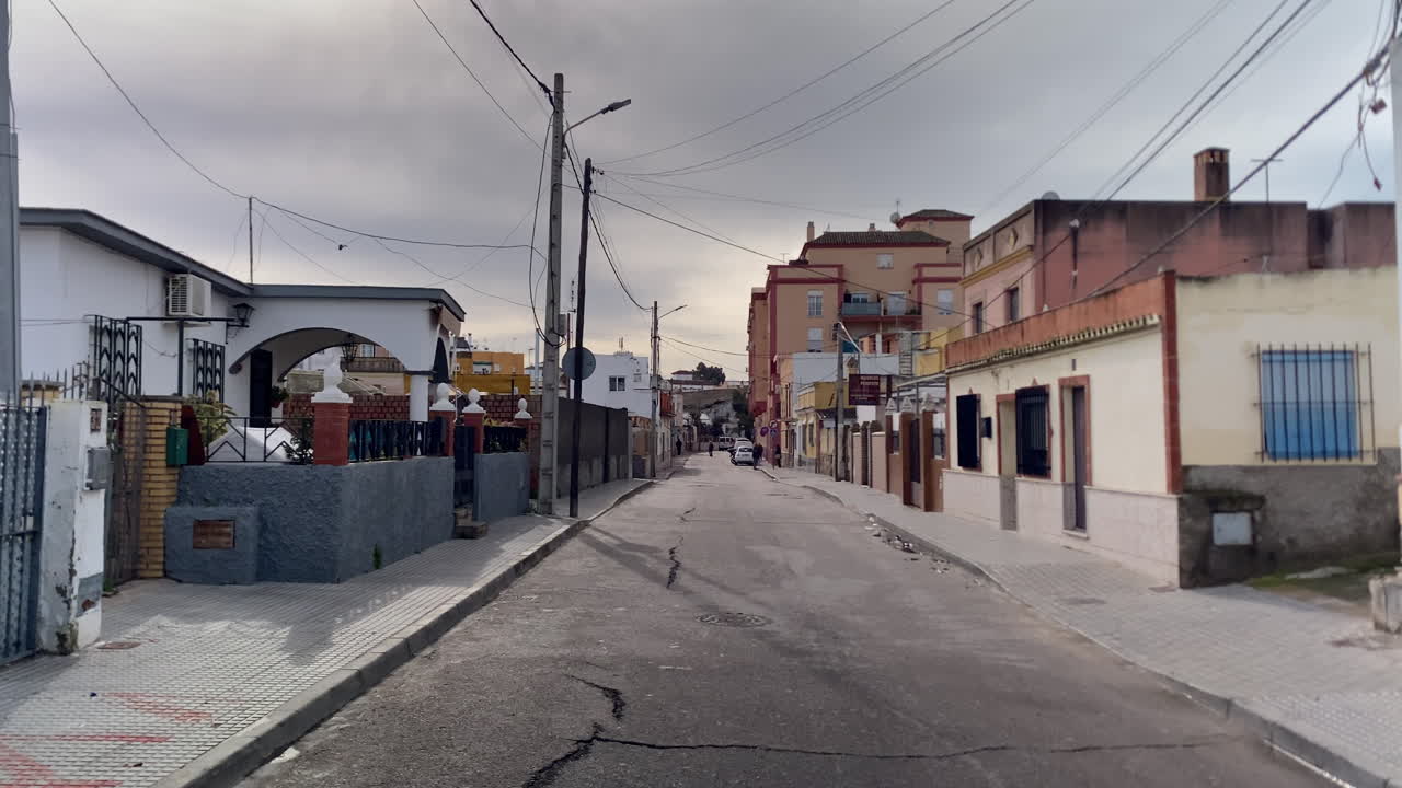 Calle residencial española en Sanlúcar de Barrameda con arquitectura local, postes de servicios y asfalto agrietado bajo un cielo nublado.