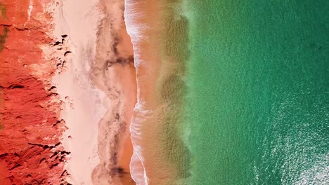 Red-cliffs-lining-sandy-beach-as-small-waves-break-on-sand,-straight-down-aerial