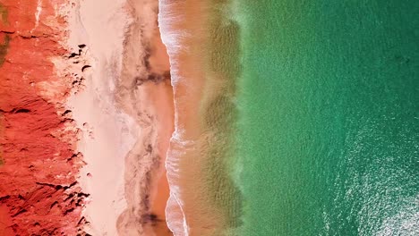 Acantilados-Rojos-Que-Bordean-La-Playa-De-Arena-Mientras-Pequeñas-Olas-Rompen-En-La-Arena,-Hacia-Abajo-Aérea