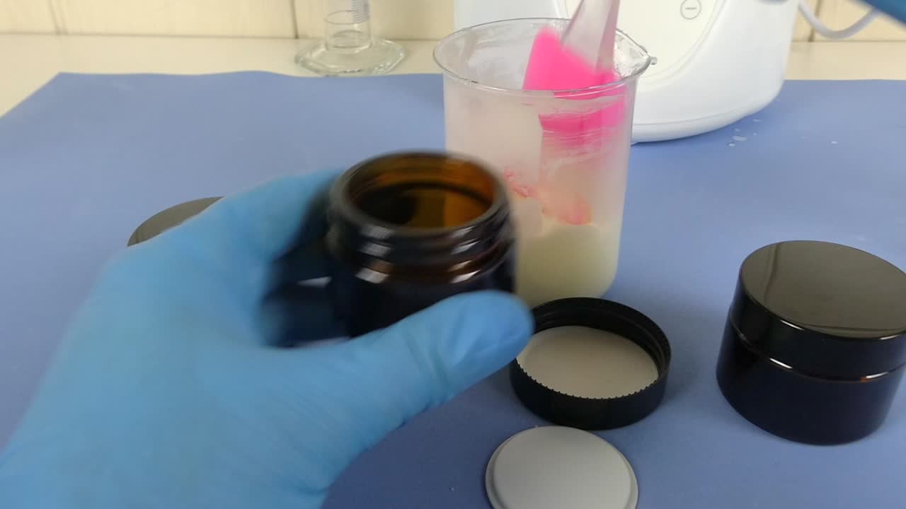 Macro view of a cosmetic laboratory scene where a gloved operator uses a spatula to carefully package a white emulsion.
