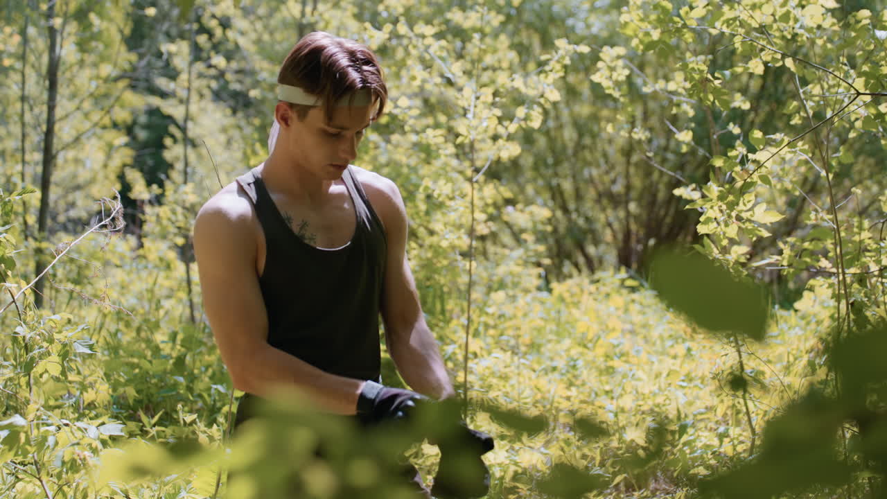 Soldier wearing headband adjusts fingerless gloves while standing amid dense forest foliage, sunlight dappling shoulders and arms, green cargo attire blending with leaves and undergrowth