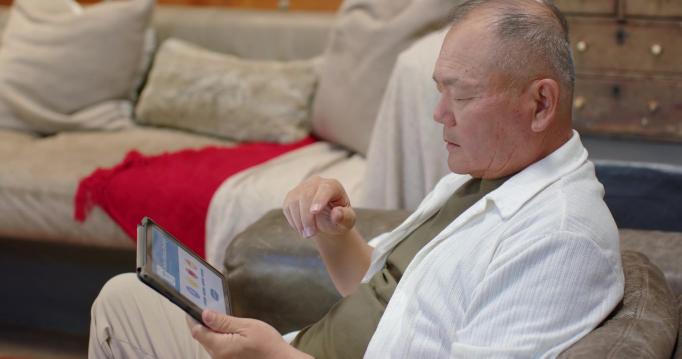 リビングルームのソファに座ったアジア系の高齢男性が、タブレットの画面を閲覧しタップしている。コピースペースあり。