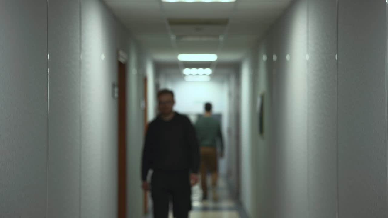 Corridoio d'ufficio con persone che camminano verso la telecamera, atmosfera da pendolarismo mattutino, illuminazione fluorescente, passaggio.