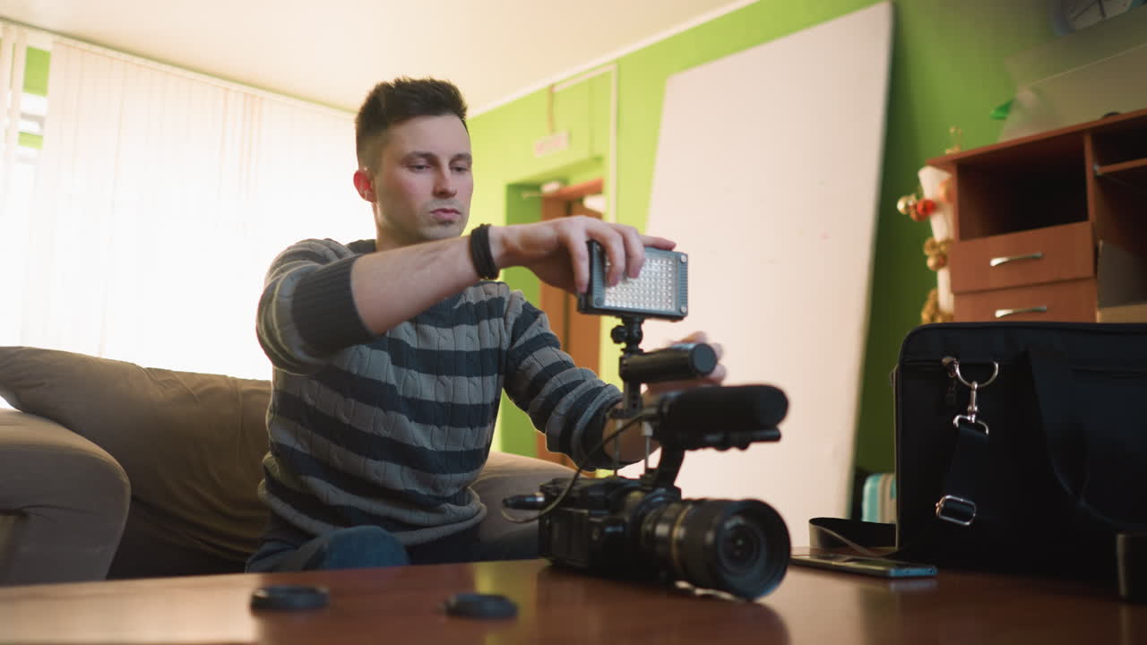 Studio guy sitting on sofa in home studio, fixing lighting , while preparing for video shoot. Camera gear with microphone, lighting, and accessories arranged on table