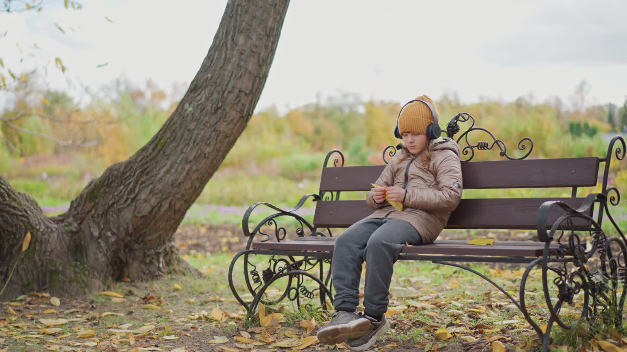 calm girl wearing mustard beanie and headphones sits on park bench under autumn trees, thoughtfully squeezing golden leaf while listening to music in crisp fall air with leaves scattered around her